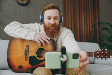 Wall Mural - Young ginger man wears casual clothes headphones play guitar use mobile cell phone sit on grey sofa couch stay at home hotel flat rest relax spend free spare time in living room indoor Lounge concept.