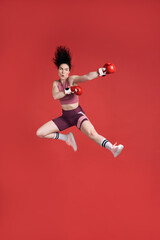 Strong sportswoman boxer fighter in red boxing gloves prepares high kick, isolated on red background