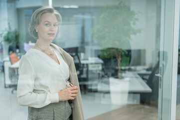 Portrait of a beautiful gray-haired caucasian woman in the office standing behind a glass wall. 