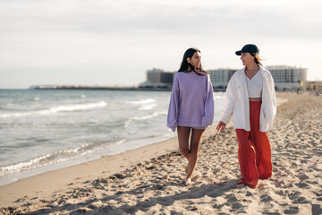 Two best friends looking at each other and having conversation while walking barefoot on sandy beach and enjoying their morning. Young women talking during relaxing morning walk on beach.