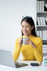 Poster - Adorable young female office worker having a coffee while working on laptop computer at the office room.