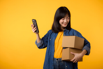 Portrait asian young woman wearing yellow t-shirt and denim shirt holding mobile phone and parcel box isolated on yellow studio background, Delivery courier and shipping service concept.