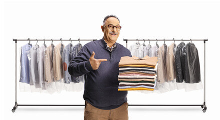 Poster - Cheerful mature man holding a pile of folded clothes and pointing in front of racks with hanging clothes