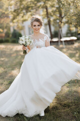 Wall Mural - Wedding portrait. A blonde bride in a white dress with a train is walking, smiling and holding a bouquet and her wedding dress. Photo session in nature. Sun rays in the photo