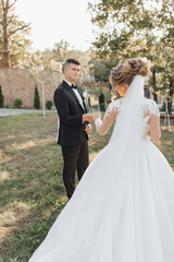 Wall Mural - Wedding portrait. A groom in a black suit and a blonde bride walk holding hands in a garden. Rear view. A white, long veil in the air. Photo session in nature. Beautiful hair and makeup