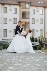 Wall Mural - Wedding portrait. A groom in a black suit and a blonde bride dance a waltz against the background of buildings. Long dress in the air. Photo session in nature. Beautiful hair and makeup