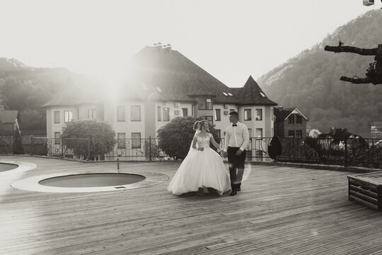 Wedding black and white portrait. The groom in a black suit and the blonde bride are walking on the background of the mountain and buildings, near the pool. Long dress in the air. Romantic photo