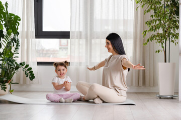 A smiling young woman is working out sitting on a fitness mat, together with a cute little daughter at home