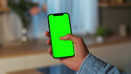 Male hand holding smartphone with green screen in vertical position. Man using mobile phone on blurred background of modern kitchen in evening. Chroma key, mockup concept. Indoors