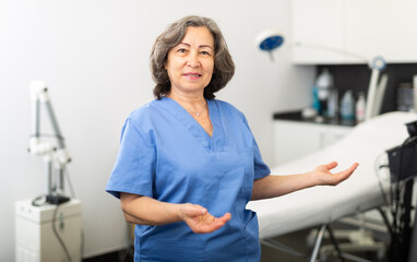 Wall Mural - Smiling experienced elderly female cosmetologist in blue overall inviting to modern aesthetic medicine office