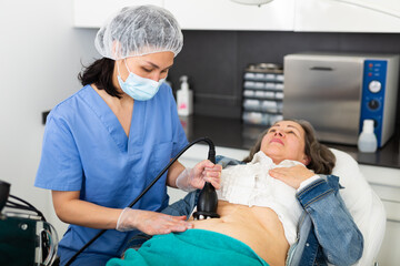 Asian woman professional cosmetologist performing abdominal skin rf lifting procedure on modern equipment for aged female patient in aesthetic medicine office