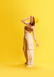 Full-length portrait of stylish young girl in trendy cargo pants, knitted top and panama standing against yellow studio background. Concept of human emotions, youth culture, fashion, lifestyle