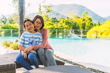 Portrait of Asian middle-aged mother and 8 or 9 years old daughter, sitting on a sunbed in the front of swimming pool of a resort, blurred background of the mountain, and empty space for text.