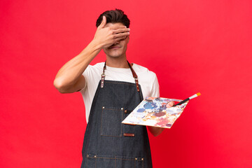 Wall Mural - Young artist caucasian man holding a palette isolated on red background covering eyes by hands. Do not want to see something