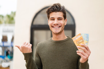 Wall Mural - Young caucasian man taking a lot of money at outdoors pointing to the side to present a product