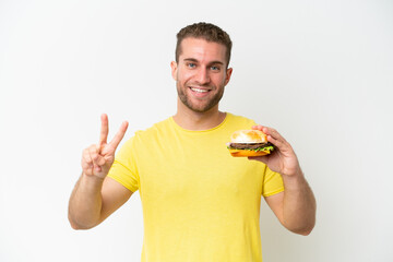 Canvas Print - Young caucasian man holding a burger isolated on white background smiling and showing victory sign