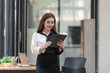 © Songsak C - Smiling young Asian businesswoman holding a digital tablet standing in an office.