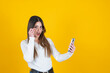 © Designerant - Surprised young woman, portrait of young caucasian surprised young woman. Shocked 20s caucasian girl looking smartphone and under glasses with open mouth. Isolated yellow orange background. Copy space