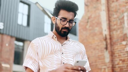 Stylish young indian man with glasses hold smartphone scrolling social media texting browsing online and looking at camera in city centre Student male relax enjoying great day outdoors 