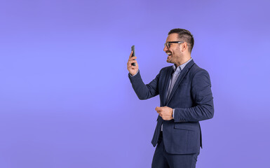 Wall Mural - Successful male professional entrepreneur laughing and talking over video call on smart phone. Young elegant businessman cheerfully discussing with client over web conference against blue background