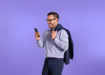 Wall Mural - Happy young businessman holding blazer and checking messages on smart phone. Male professional entrepreneur smiling and scrolling social media while standing isolated against blue background