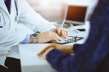 Wall Mural - A doctor is talking to his patient, while sitting together at the desk in the sunny cabinet in a hospital. Physician using clipboard for filling up medication history records. Perfect medical service 