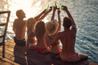 © luckybusiness - Rear view shot of group of young people sitting on wooden jetty by water, cheering with drinks. Couples on vacation together. Holiday, togetherness, lifestyle concept.