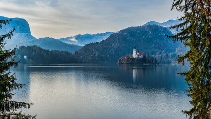 Wall Mural - Panorama shot of lake Bled Pilgrimage Church island and the mountain range framed bewteen trees during winter, Slovenia