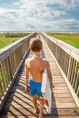 Wall Mural - Young boy in swim trunks walking on a bridge toward the beach while carrying his boogie board