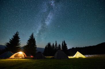 night camping in mountains under starry sky. tourist tents in campsite near burning campfire under b