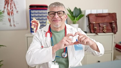 Poster - Middle age grey-haired man doctor smiling doing heart gesture with hands at clinic