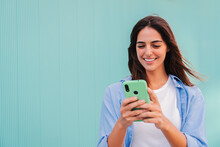 Smiling Happy Teenage Woman Using Her Smartphone And Chatting Online ,or Browsing On Internet At Blue Teal Wall Background. Young Caucasian College Student Lady Watching Funny Videos With Mobile Phone