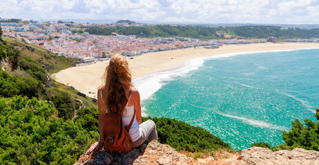 Wall Mural - woman tourist sitting on cliff and enjoying panoramic view of atlantic ocean, nazare beach in portug