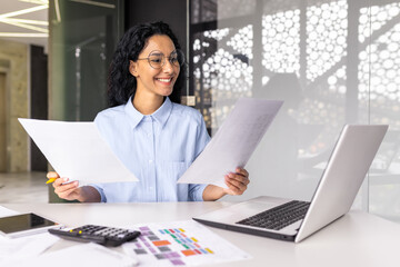 Wall Mural - latin american businesswoman working inside office with documents and laptop, worker paperwork calculates financial indicators smiling and happy with success and results of achievement and work.