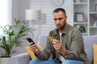 Worried young hispanic man sitting on sofa at home and looking at credit card upset. Holds the phone in his hand, cannot transfer money, make a payment, the account is blocked, bankruptcy.