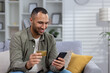 © Tetiana - Smiling young African American man sitting on sofa at home and using credit card and phone, checking account, making money transfer, online shopping
