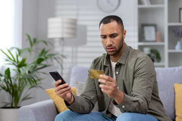 worried young hispanic man sitting on sofa at home and looking at credit card upset. holds the phone