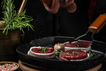 Wall Mural - Chef cooking steak on grill pan. male hands holding Beef medallions wrapped in bacon with rosemary and spices