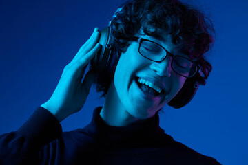 Wall Mural - Man wearing headphones listening to music, dancing and singing open mouth smile with glasses, hipster lifestyle, portrait blue background mixed neon light, fashion style and trends, copy space