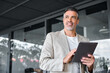 © insta_photos - Smiling mid aged business man wearing suit standing outside office holding digital tablet. Mature businessman professional holding fintech device looking away thinking or new business ideas solutions.