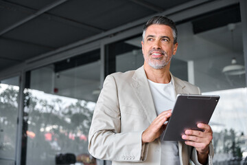 Smiling mid aged business man wearing suit standing outside office holding digital tablet. Mature businessman professional holding fintech device looking away thinking or new business ideas solutions.