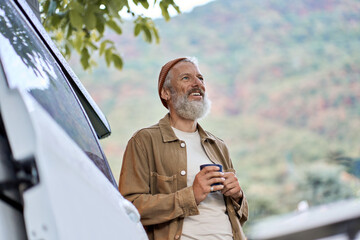 Active old happy hipster man standing near rv camper van on vacation. Mature traveler looking away enjoying view, holding drinking coffee waking up in the morning in camping tourism nature park.