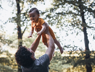 Caucasian young dad throws up his little daughter in a yellow overalls, looking into daddy's eyes