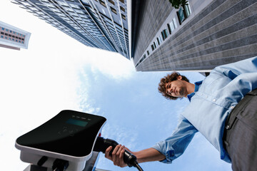 Fisheye view suit-clad progressive businessman at charging station for electric vehicle at modern city with the residential apartment condo building in the background. Idea for eco-friendly car.