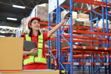 Warehouse workers woman with hardhats and reflective jackets using computer and pointing something with walkie talkie radio while controlling stock inventory in retail warehouse logistics center