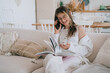 © Iona - Cheerful caucasian young girl in white casual clothes sitting on couch with book talks by phone smiles eyes closed. Teen girl laughing during conversation with friend.
