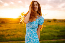 Young Woman In Stylish Summer Dress Feeling Free In The Field With Flowers At Sunset. Nature, Fashion, Vacation And Lifestyle