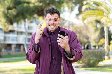 Wall Mural - Young caucasian man at outdoors using mobile phone with fingers crossing