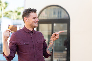 Wall Mural - Young caucasian man holding a wallet at outdoors pointing to the side to present a product