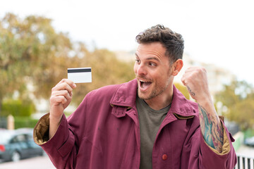 Sticker - Young caucasian man holding a credit card at outdoors celebrating a victory